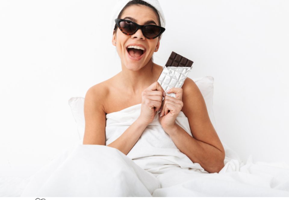 picture of a happy woman in a towel wearing sunglasses eating chocolate